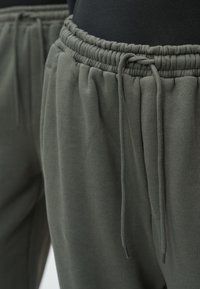 Sötétzöld szabadidőnadrág, amely elasztikus derékrésszel és állítható húzózsinórral rendelkezik, puha tapintású, oldalán ráncolt részletekkel a kényelmes illeszkedés érdekében.