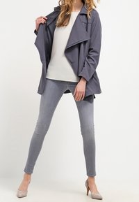 Szary, lekki płaszcz trencz z otwartym przodem, długimi rękawami i luźnym krojem, zestawiony z białą bluzką i szarymi obcisłymi dżinsami.