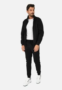 Giacca nera con zip indossata sopra una maglietta bianca, abbinata a pantaloni della tuta neri e sneakers bianche. La texture è liscia, con polsini aderenti.