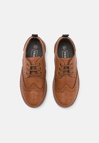Bruna läderskor med brogue-detaljer, rund tå, stuckna kanter och tunna snören. Utrustade med en svart inredning och en hälflik.