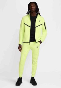 Chaqueta con cremallera de color amarillo neón con acentos en negro, combinada con pantalones jogger a juego. Tejido texturizado y diseño deportivo, con un bolsillo lateral y un logo.