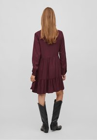 Bordeaux klänning med långa ärmar, vågig nederkant och lätt material, i kombination med knähöga svarta stövlar. Visas från baksidan.