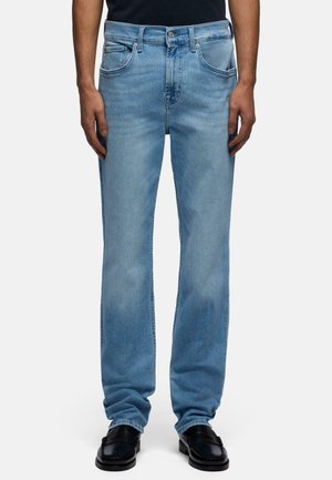 Hellblaue Jeans mit geradem Bein, getragen von einer Person mit schwarzen Schuhen, dargestellt von der Taille bis zu den Füßen vor einfachem Hintergrund.