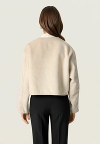 Cropped beige jacket made of soft material with long sleeves, rounded neckline, and subtle stitching details; paired with black pants.