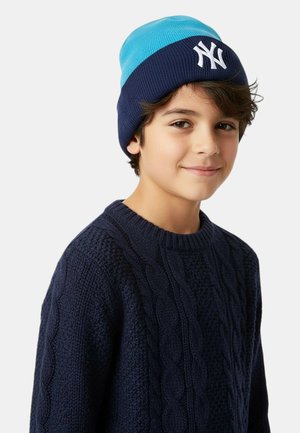 Ragazzo che indossa un maglione a trecce blu e un berretto blu e azzurro con il logo bianco dei New York Yankees.