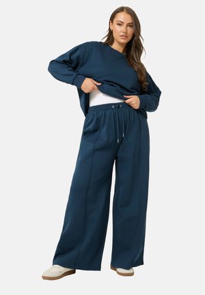 Frau hebt einen navyblauen, übergroßen Pullover hoch, trägt passende weite Hosen und weiße Turnschuhe, steht vor einfarbigem Hintergrund.