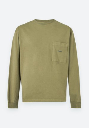 Jeune homme portant une chemise à manches longues vert olive avec une poche poitrine, associée à un pantalon en jean foncé et ample, debout devant un fond uni.