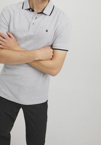 Grå polo tröja med strukturerad yta, svart krage och ärmslutar, treknapps stängning och en liten logotyp på vänster bröst.