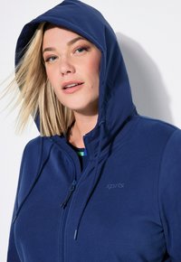 Giacca a vento blu navy con zip completa, caratterizzata da un tessuto testurizzato, cordini regolabili e un logo discreto sul petto.