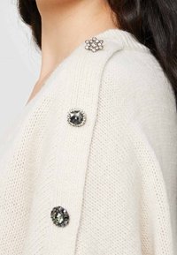 Kremowy sweter dzianinowy z dekoracyjnymi guzikami: jeden okrągły czarny kamień, jeden okrągły metalowy akcent oraz jeden kwiatowy wzór z przezroczystymi kamieniami.