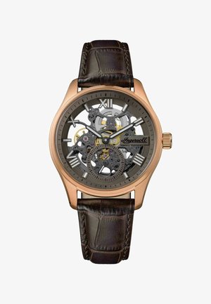 Orologio scheletro con cassa in bronzo, quadrante grigio e movimento esposto. Il cinturino in pelle marrone presenta una finitura testurizzata. Numeri romani alle ore 12 e 6.
