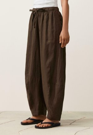 Osoba stojaca na dláždenej podlahe, ktorá má na sebe voľné tmavohnedé ľanové nohavice s elastickým pásom a šnúrkou, biely top a čierne žabky.