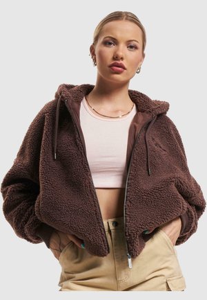 Brun fleecehoodie med mjuk textur, framdragkedja och snörning i huvan, bärs över en ljusrosa crop top och beige byxor.