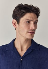 Narančasta košulja s kratkim rukavima napravljen od teksturiranog tkanja, s klasičnim ovratnikom i zatvaranjem na gumb. Suptilni uzorak dodaje dubinu dizajnu.