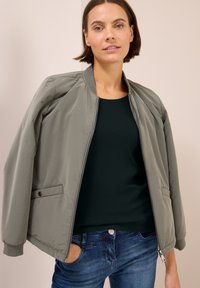 Frau in olivgrüner Bomberjacke über schwarzem Oberteil und blauen Jeans, steht mit einer Hand in der Tasche vor einem schlichten Hintergrund.