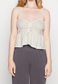 Ljusbeige kort topp med tunna justerbara band, texturerat tyg, sammanknuten detalj fram, och en utsvängd fåll. Matchad med grå byxor.