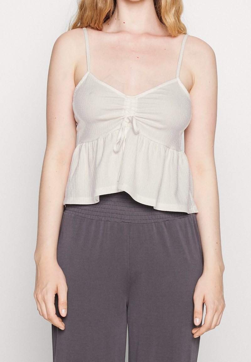 Ljusbeige kort topp med tunna justerbara band, texturerat tyg, sammanknuten detalj fram, och en utsvängd fåll. Matchad med grå byxor.