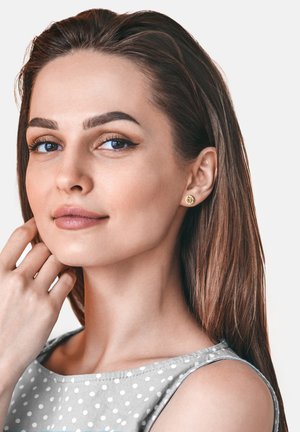 Orecchini a bottone in oro con un motivo floreale, indossati da una donna con lunghi capelli castani, in una maglietta a pois grigi, su uno sfondo neutro.