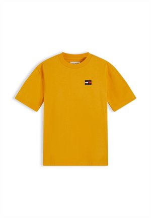 Enfant souriant aux cheveux bouclés portant un t-shirt jaune à manches courtes, un pantalon ample foncé, des baskets blanches, debout les mains dans les poches.