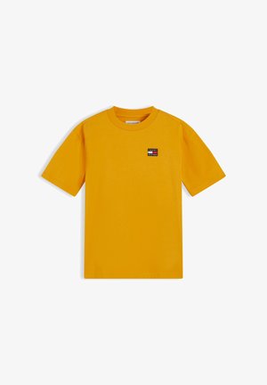Enfant souriant aux cheveux bouclés portant un t-shirt jaune à manches courtes, un pantalon ample foncé, des baskets blanches, debout les mains dans les poches.