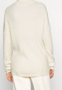 Strickpullover in cremefarben mit gerippter Struktur, lockerer Passform und überschnittenen Schultern, ausgestattet mit langen Ärmeln und einem geschwungenen Saum.