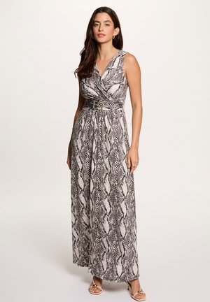 Vrouw draagt een mouwloze maxi-jurk met ceintuur en zwart-witte slangenprint, gecombineerd met beige open teen hakken, staand tegen een effen achtergrond.