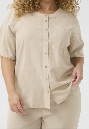 Kvinde med krøllet hår iført en beige kortærmet skjorte med knapper og matchende bukser, der holder skjortens kant med den ene hånd.