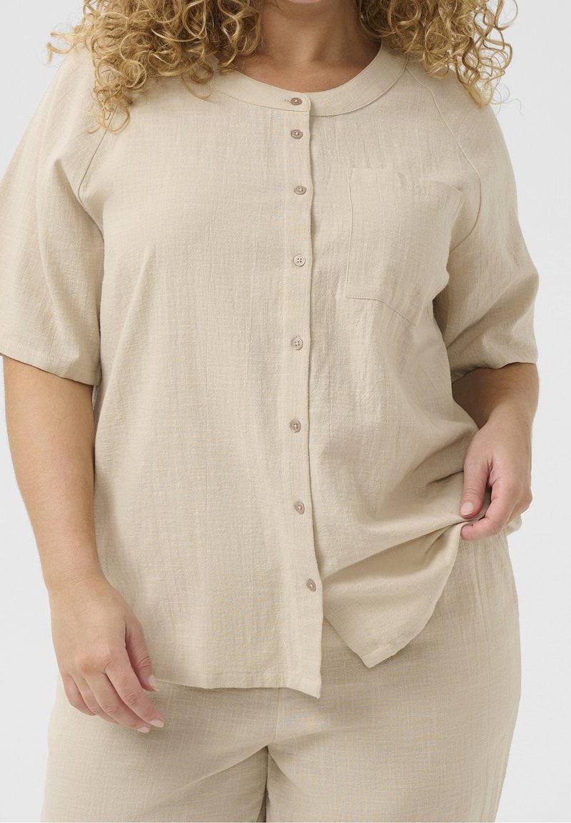 Femme aux cheveux bouclés portant une chemise beige à manches courtes boutonnée et un pantalon assorti, tenant l'ourlet de la chemise d'une main.