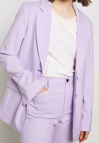 Levandulový blazer s klopovými límci a dvěma knoflíky, spárovaný se světle růžovým tričkem s tkaninovým vzorem a odpovídajícími levandulovými kalhotami.