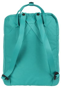 Mochila de tela en color azul verdoso con superficie texturizada, correas ajustables y asa superior. Presenta un diseño cuadrado minimalista y costuras reforzadas.