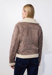 Brun semsket skinnjakke med pelskrage og mansjetter. Har en tettsittende design og justeringsremmer på siden. Bæres med mørk denim.