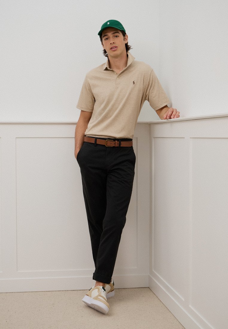 Polo beige, pantalon noir, casquette verte et baskets beige-blanc, avec une ceinture brune. L'ensemble présente une coupe décontractée et des éléments de design classiques.
