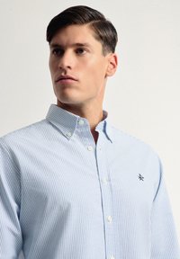 Camisa de botones de rayas azul claro con cuello, con botones blancos y un pequeño logo bordado en el pecho. Textura de algodón suave.