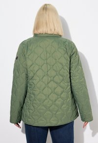 Giacca trapuntata verde con una superficie texture, colletto rotondo e maniche lunghe. Presenta una toppa nera con logo sul braccio sinistro.