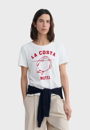 Woman wearing white T-shirt with red "LA COSTA MOTEL" text and seal graphic, beige pants, and a dark sweater tied around her waist.