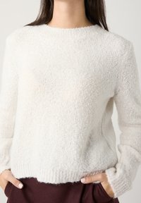 Pull blanc tricoté avec une texture duveteuse, col rond, et poignets et ourlet côtelés. Porté avec un pantalon brun foncé.