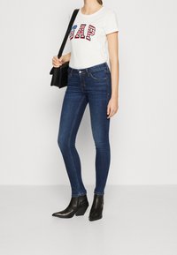Camiseta blanca de manga corta con letras "GAP" en rojo y azul, combinada con jeans skinny azul oscuro y botines negros de tacón.