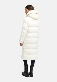 Long, puffy white coat with an attached hood. Features a quilted design and a mid-calf length. Worn over black pants and chunky black boots.