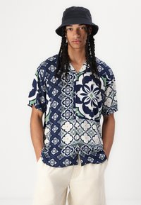 Kurzärmliges Button-Up-Hemd mit einem floralem Muster in Marineblau, Grün und Weiß; das Modell trägt einen schwarzen Bucket Hat und beigen Cordshorts.