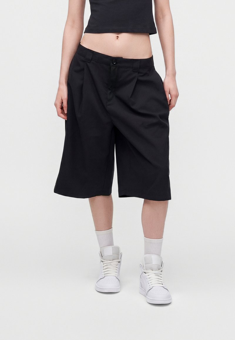 Personne portant un short large noir au-dessus du genou, des baskets montantes blanches, des chaussettes blanches et un crop top noir sur fond blanc.