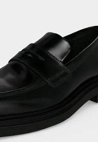 Mocassin en cuir noir poli avec un petit talon et une bride sur le devant supérieur avec un détail découpé, présenté sur fond blanc.