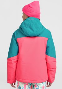 Gevoerde jas met een teal bovenkant en felroze onderkant. Beschikt over een capuchon en een zachte textuur. Opvallend logo op de achterkant. Multikleurige gepatternde broek.