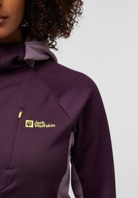 Paarse fleece hoodie met grijze zijpanelen, voorzien van een geel logo op de borst en een voorste ritszak, textuurstof en hoge kraag.