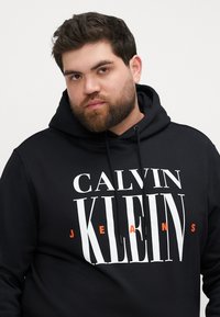 Hanorac negru cu buzunar cangur, glugă cu șnur și text mare alb "CALVIN KLEIN" cu "JEANS" în portocaliu. Textură de material neted.