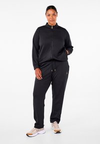 Černý zip-up sportovní oblek s vysokým límcem, bočními kapsami a kalhotami na stahování. V kombinaci se světlými sportovními botami s hnědými akcenty.