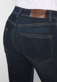 Jeans de mezclilla oscura con cintura alta, que cuentan con un parche de cuero marrón de la marca, costuras amarillas y dos bolsillos traseros.