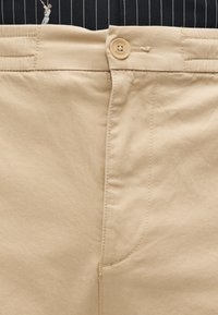 Chinos kaki à texture lisse, avec fermeture éclair et bouton, et poches cousues. Logo brodé visible près de la taille.