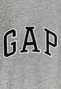 Tela de sudadera gris con un logo "GAP" bordado en negro contorneado en blanco. El diseño es sencillo y la textura parece suave.