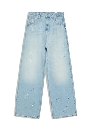 Homme portant un jean oversized, un t-shirt blanc, des lunettes de soleil et des sandales, debout sur un sentier de jardin près de statues classiques en plein jour.