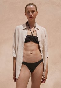 Svetlo bež lanena srajca nad črnim bikinijem z nabranimi detajli in usklajenimi bikini hlačkami. Gladek, teksturiran material ob koži.